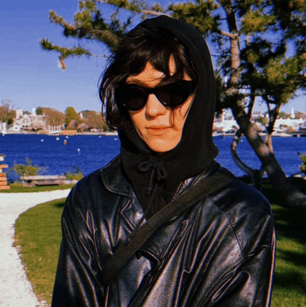 a woman in all black with dark sunglasses, standing in front of a body of water.